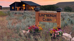 Een hutje, genaamd 'Lindbergh Cabin', met een bord ervoor, omringd door bloemen en een ondergaande zon op de achtergrond.