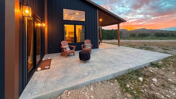 Een modern gebouw met een overdekte patio, twee Adirondack-stoelen en een uitzicht op de ondergaande zon boven een heuvelachtig landschap.