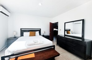 A hotel room with a bed, bedside tables, a dresser, a mirror, and an air conditioning unit.