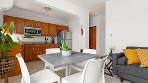 A modern kitchen with wooden cabinets, a stainless steel refrigerator, and a microwave. There is a dining table with white chairs and a potted plant. A gray sofa with a mustard pillow is in the living area. - Vacation Getaway in Ocean Park With Parking (San Juan)