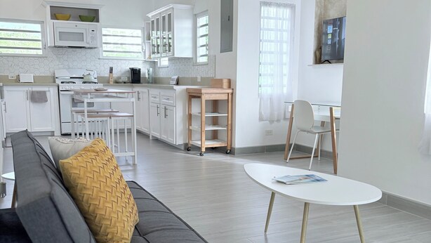 A modern kitchen with white cabinets, a microwave, and a stove. There is a dining area with a gray sofa and a white table.