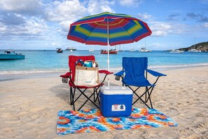 A beach scene with two beach chairs, a colorful umbrella, and a cooler on a towel.