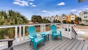 Une terrasse en bois, agrémentée de chaises Adirondack turquoise et d’une table, offre une vue sur une rangée de maisons de ville aux couleurs vives.