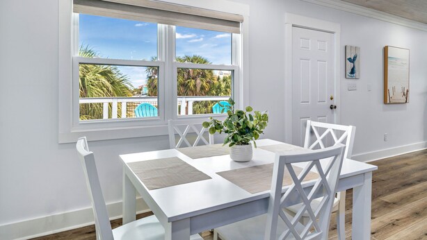 Un coin repas avec une table et des chaises blanches, une plante en pot et une vue sur des palmiers à travers de grandes fenêtres.