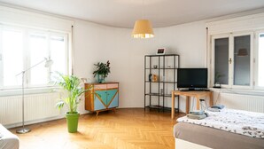 Una habitación con piso de madera, una planta en maceta verde, un mueble de almacenamiento colorido, un soporte negro para televisor y una cama con colcha estampada.