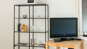 Una habitación con piso de madera, un televisor negro sobre una mesa de madera, una estantería metálica negra con varios objetos y una lámpara colgante beige de gran tamaño.