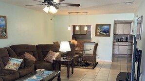 A living room with a sofa, coffee table, and a TV stand. There is a dining area with chairs and a table. The kitchen is visible in the background with cabinets and a countertop.