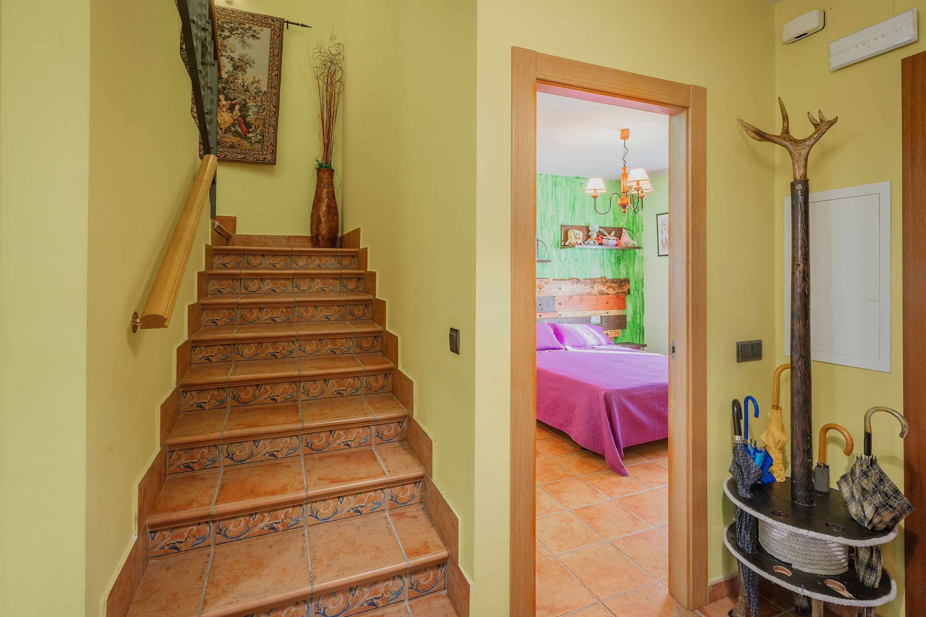 Un escalier orné de balustrades travaillées, un couloir menant à une pièce avec un lit et un mur vert, et un porte-parapluie décoratif.