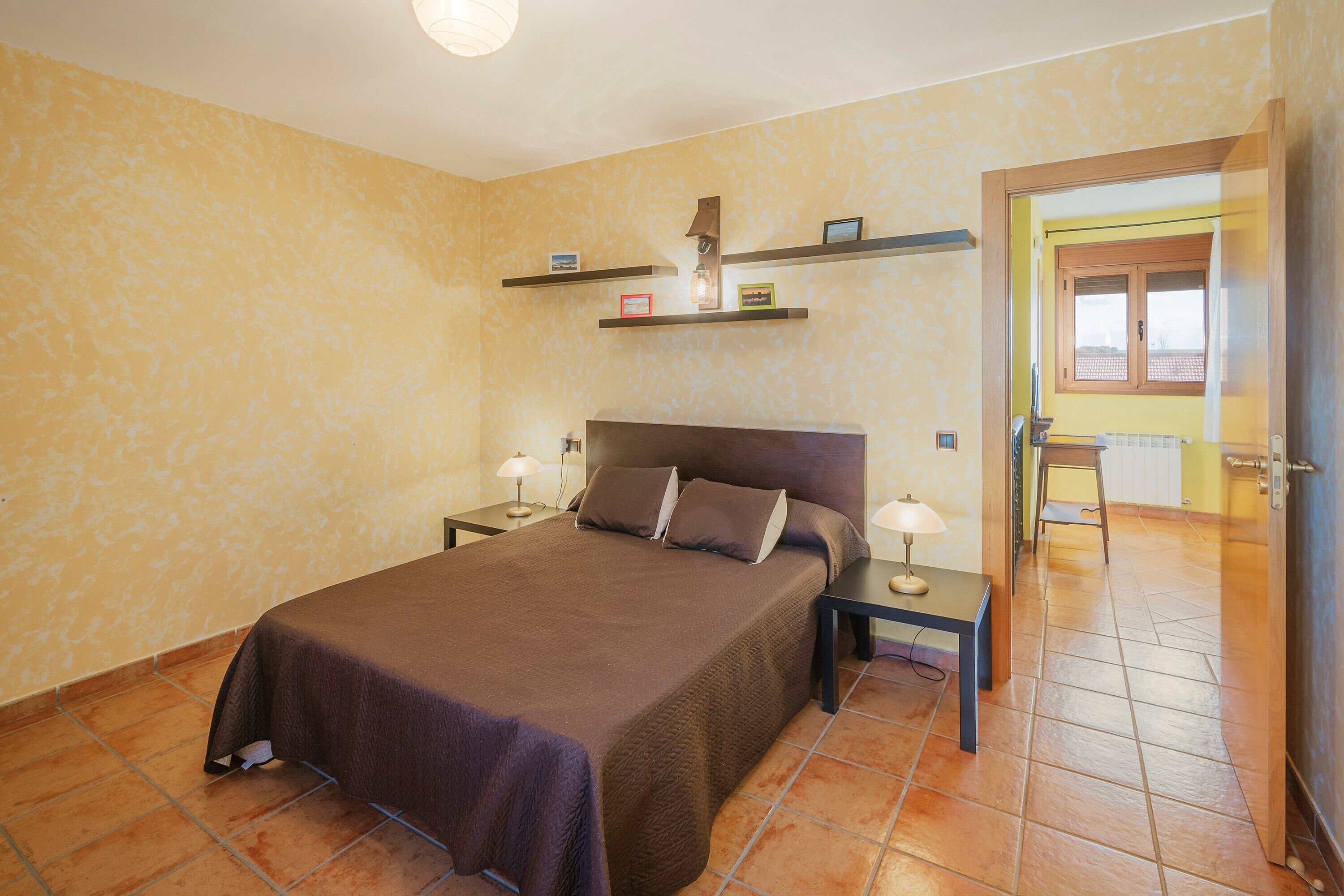 Een slaapkamer met een bed, nachtkastjes, een lamp, een plank met spullen en een deur naar een andere kamer.