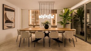 A modern dining area with a wooden table, six chairs, a hanging light fixture, and a glass cabinet with glassware.