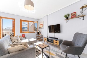 A modern living room with a sofa, armchair, coffee table, TV, and shelves with plants.
