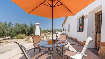 An outdoor patio with a round table and chairs, an orange umbrella, and a white building with a door and window.