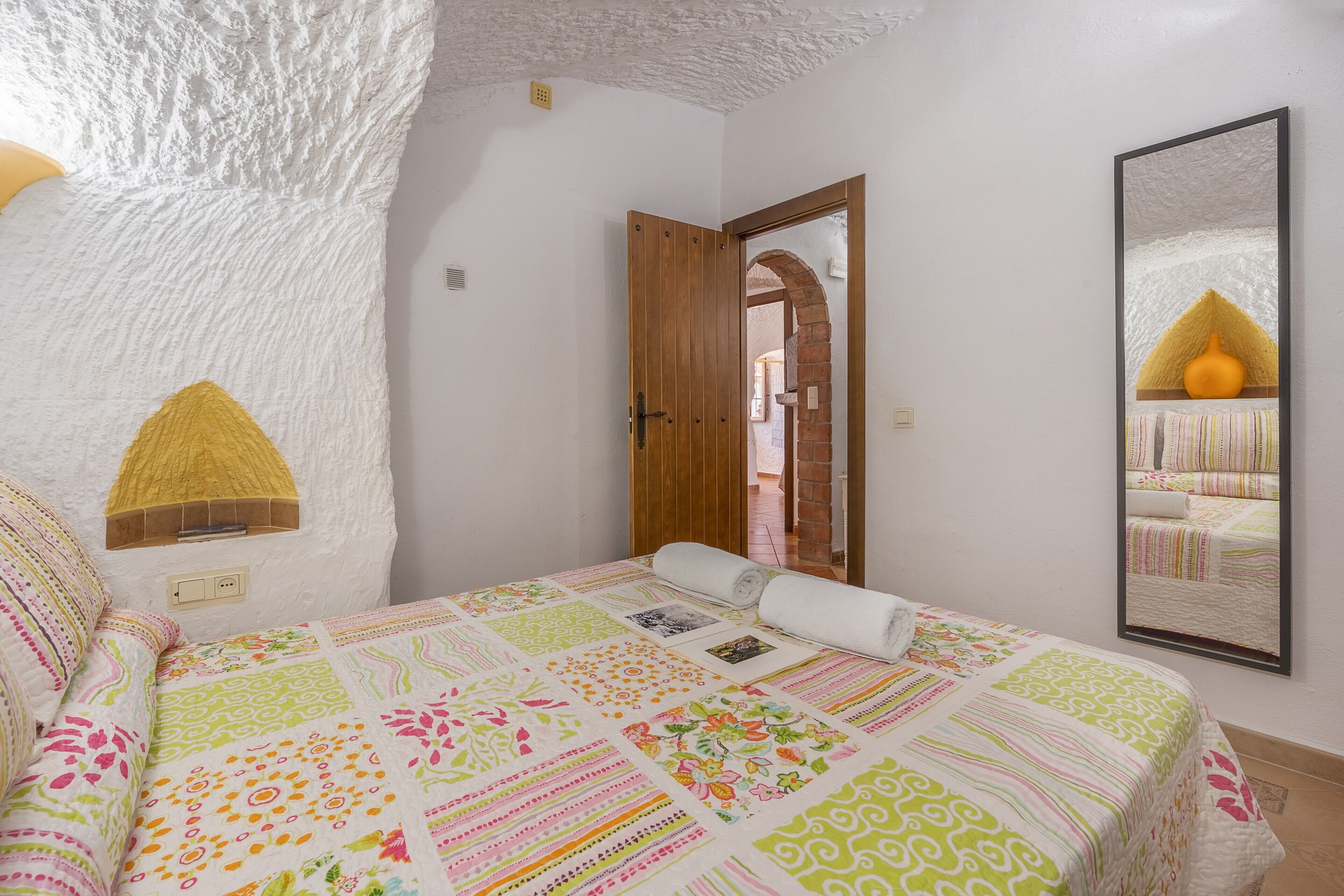 A bedroom with a patterned bedspread, a wooden door, and a mirror on the wall.