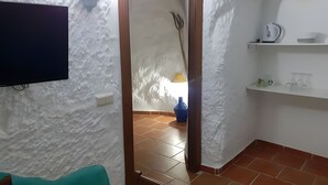 Una habitación con pared blanca con textura, un televisor de pantalla plana montado en la pared, una puerta de madera, piso de baldosas y un estante con algunos objetos.