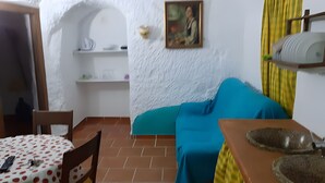 Una habitación con piso de baldosas, una cama azul, una mesa de madera con mantel estampado de lunares, una silla, una pared blanca con un cuadro y una cortina amarilla.