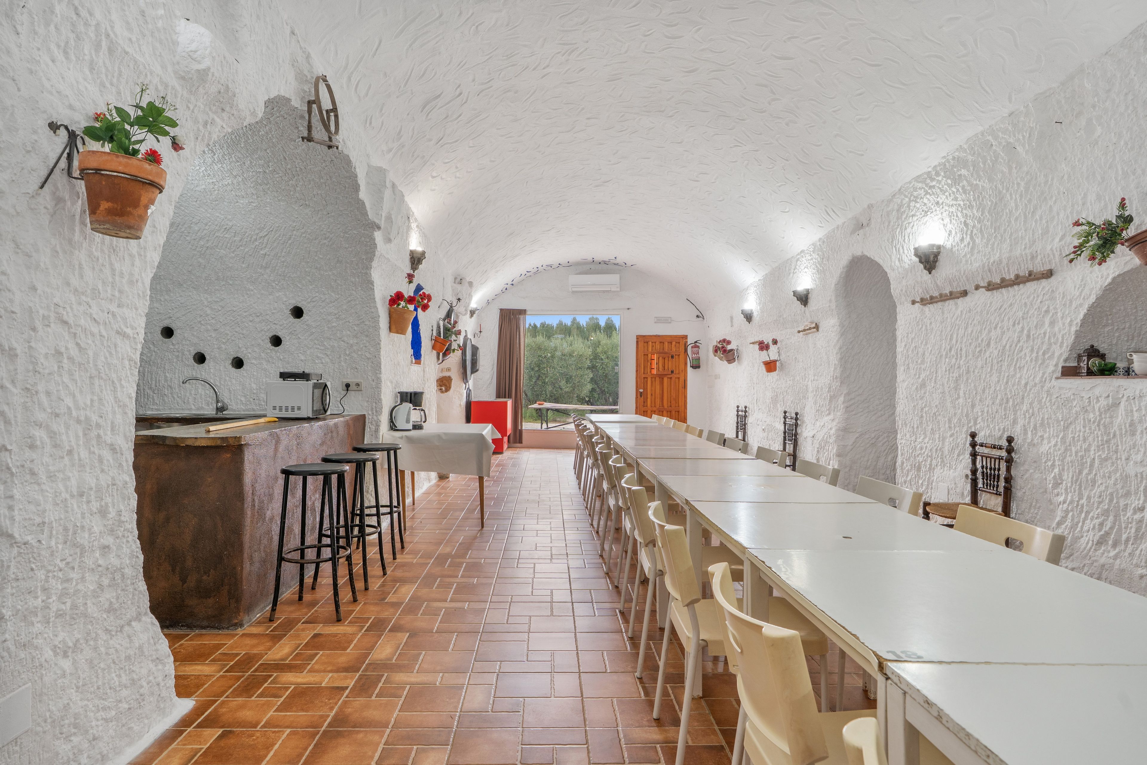 An interior with a long dining table, chairs, a kitchen area, and a view through an open door.