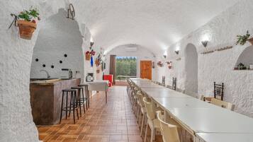 An interior with a long dining table, chairs, a kitchen area, and a view through an open door.