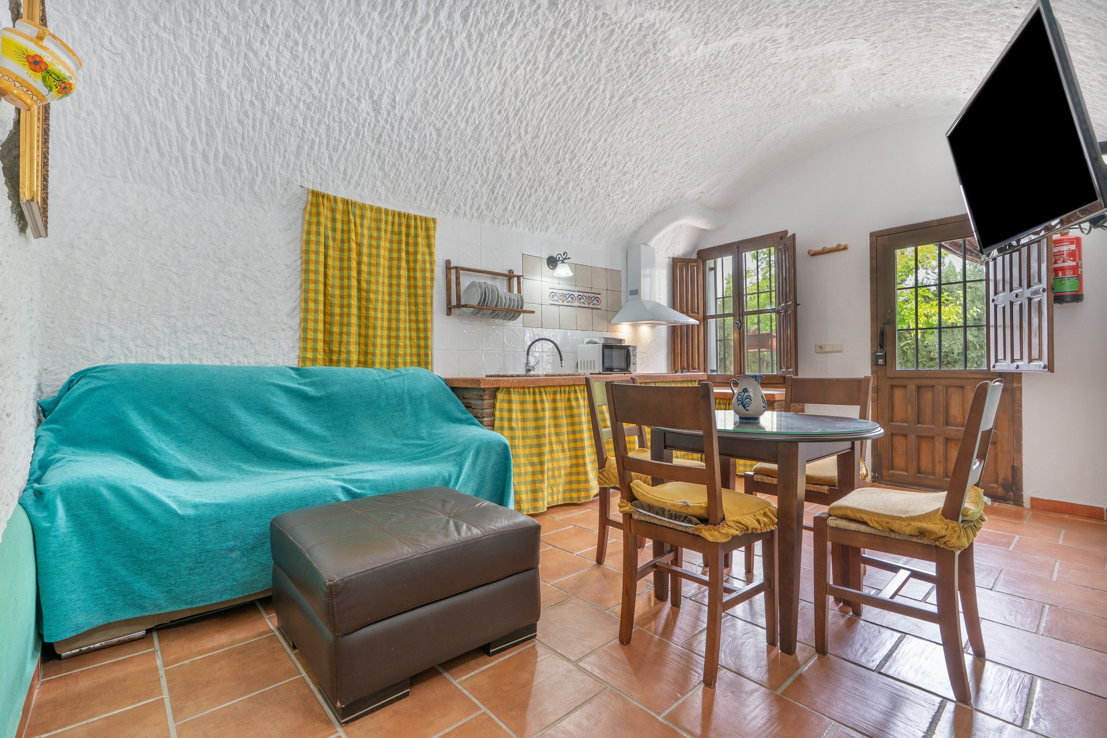 Una sala de estar acogedora con un sofá, un reposapiés y sillones, una zona de cocina con fregadero y microondas, y un televisor montado en la pared.