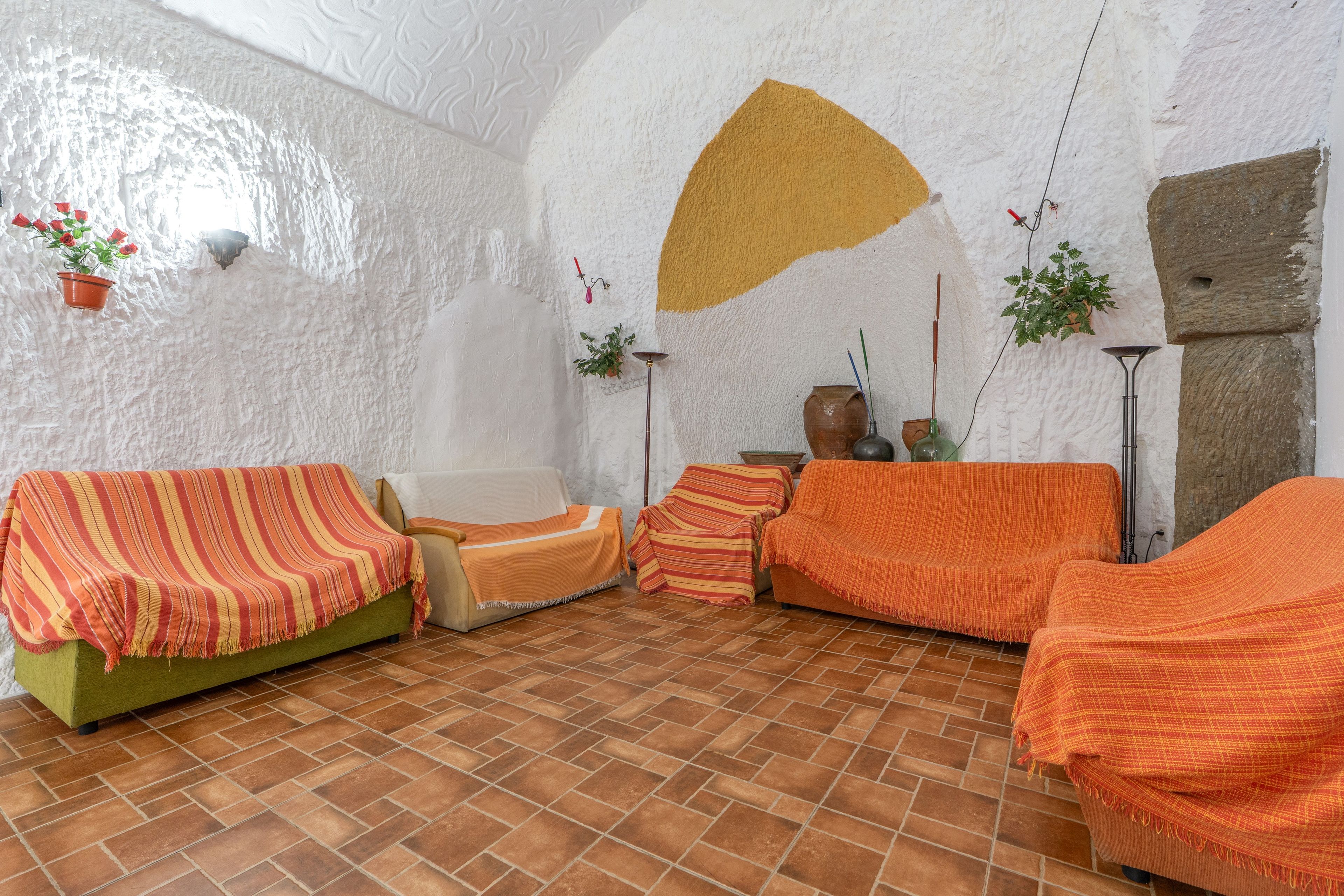 A room with a rustic aesthetic, featuring a green sofa, a brown sofa, and a patterned orange sofa. There are potted plants on the walls and a large, textured wall.