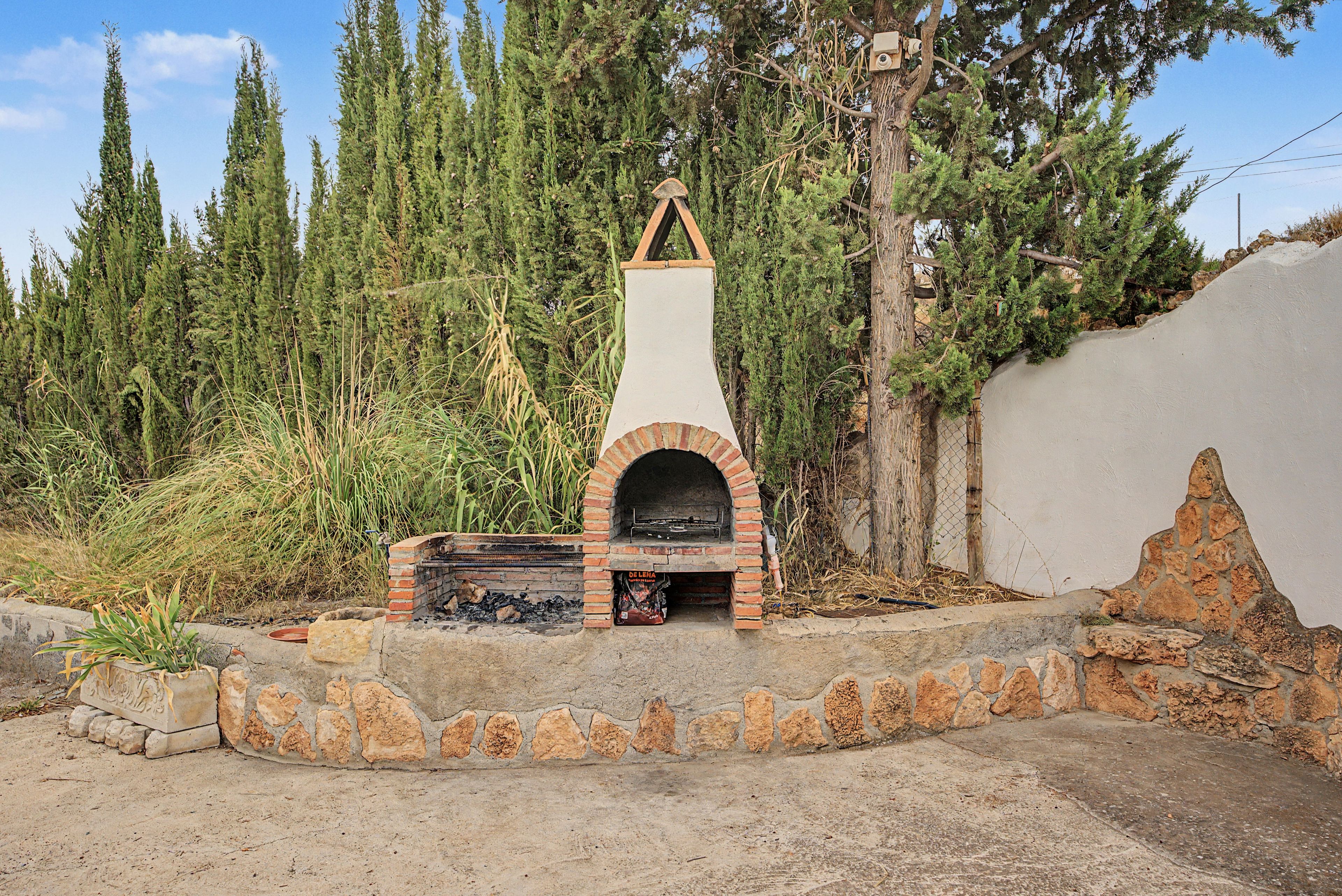 An outdoor brick oven with a chimney, surrounded by a natural landscape with tall trees and a stone wall.