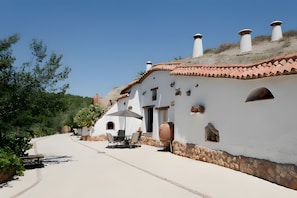 Un bâtiment blanc avec des tuiles en terre cuite sur le toit, des murs en pierre et une terrasse aménagée avec du mobilier d’extérieur.