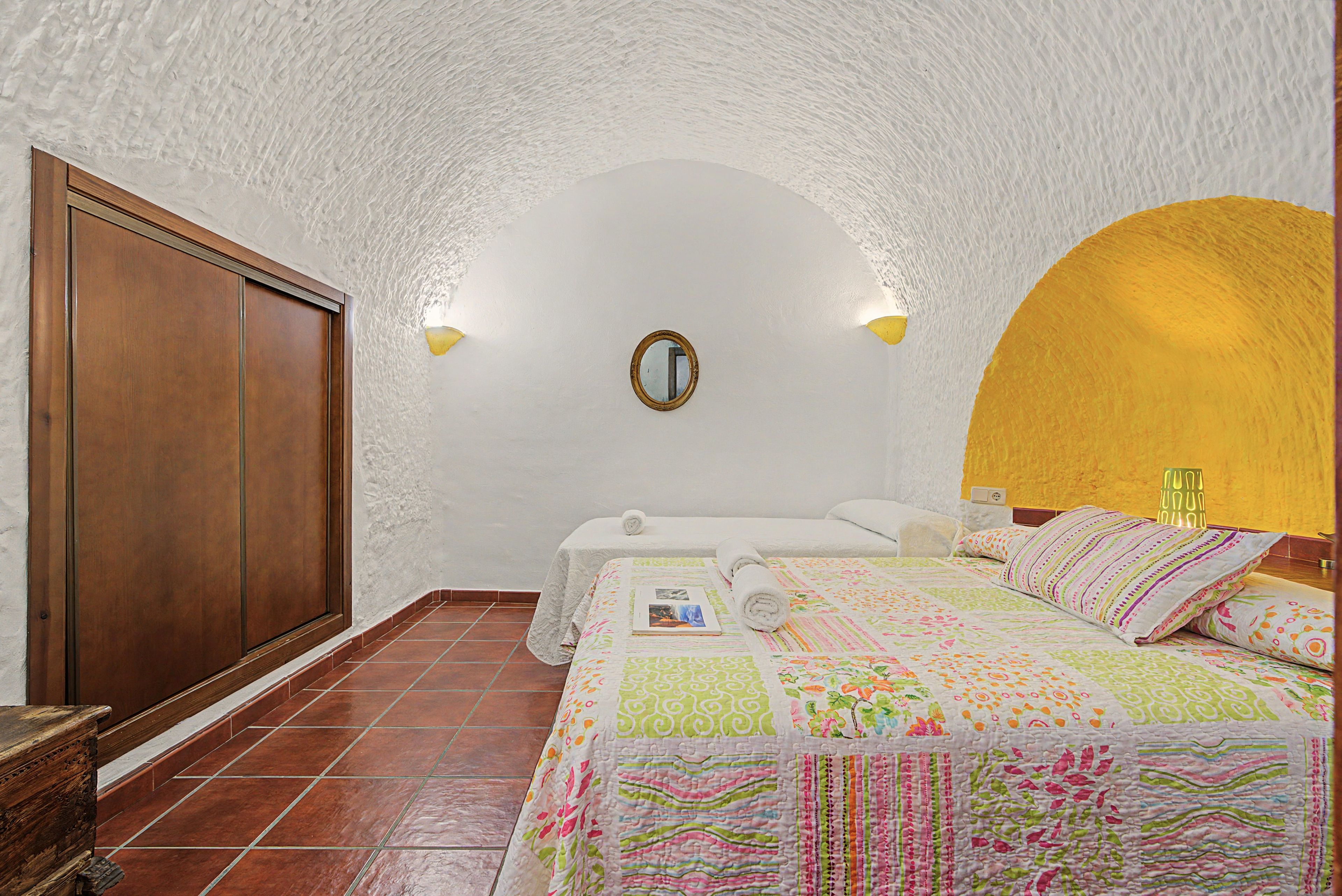 A bedroom with a wooden wardrobe, a bed with a colorful quilt, a mirror on the wall, and a yellow accent wall.