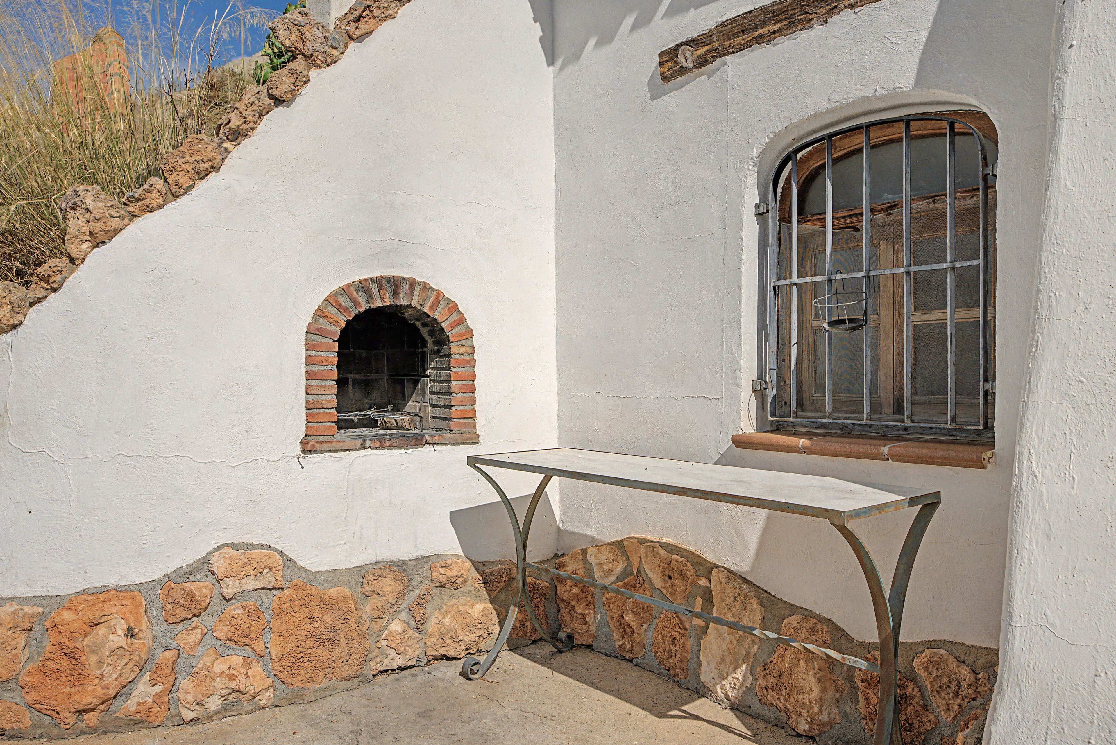 Un edificio blanco con base de piedra, una pequeña ventana en arco y una mesa de metal al frente.