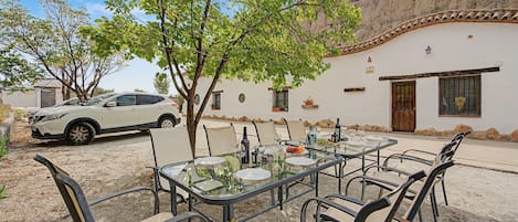 An outdoor dining area with a table set for four, surrounded by chairs, in front of a building with a white facade and a tiled roof.