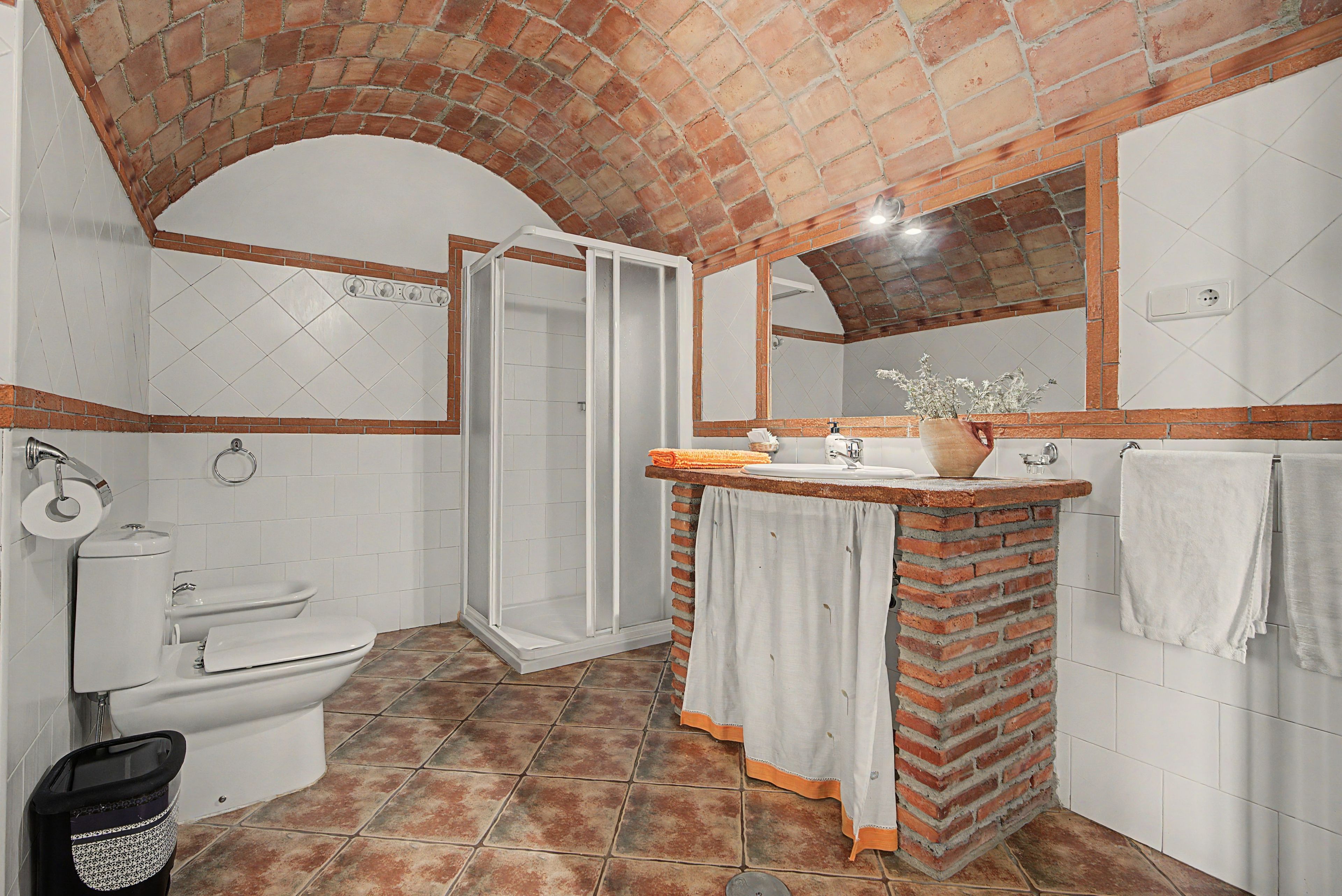 Een badkamer met douche, toilet en wastafel, met een bakstenen boog en betegelde wanden.