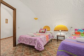A cozy bedroom with a sloped ceiling, a bed with a colorful quilt, a bedside table with a lamp, and a small window with a yellow light fixture. - Country House 'El Olivo' with Shared Pool, Wi-Fi and Air Conditioning (Benalúa)