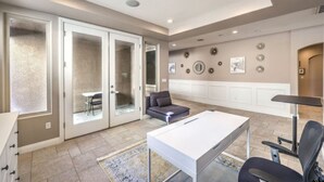 A spacious living area with a large white table, a sofa, and a chair. There are sliding glass doors leading to a balcony, and a wall with decorative plates and a clock. - Copper View-Modern 4BR, Casita, Green Valley (Henderson)