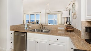 A kitchen with granite countertops, white cabinets, a stainless steel dishwasher, and a view of the ocean through large windows. - Treasure Island 312 | Elite 3BR View (Panama City)