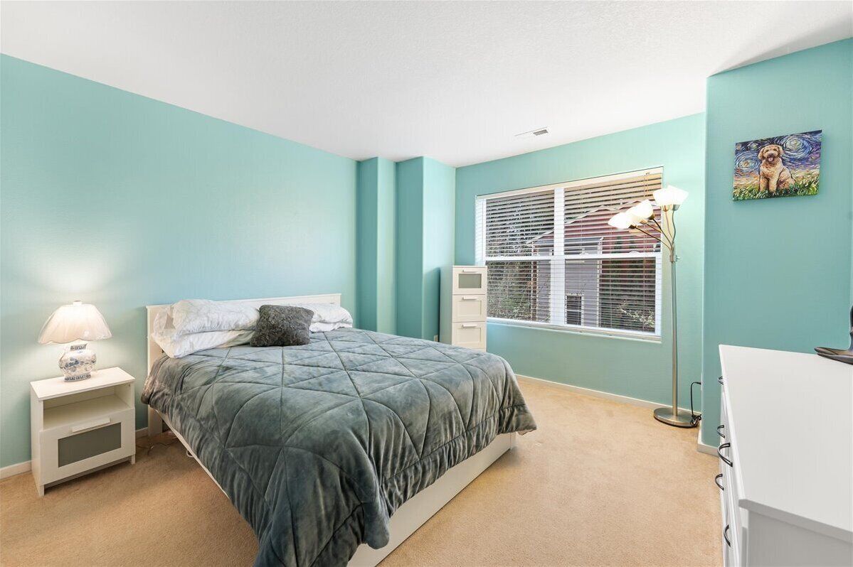 Une chambre avec des murs de couleur turquoise, un lit recouvert d’une couette grise, une table de chevet blanche avec une lampe, une fenêtre avec des stores et un tableau accroché au mur.