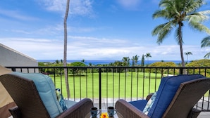 A balcony with wicker furniture and a table, overlooking a well-maintained lawn and palm trees, with a clear view of the ocean. - 🏖️Ocean View Kona Condo w/ AC | Golf Course + Lanai (Kailua Kona)