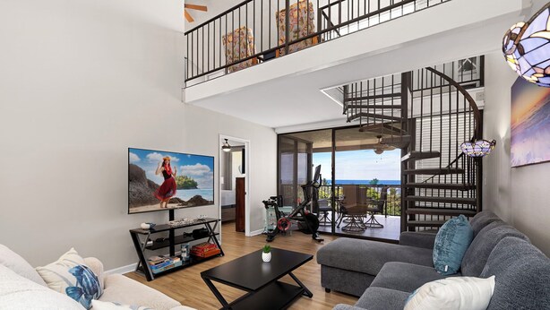 A spacious living area with a grey sofa, a black coffee table, and a large painting of a woman on a rock. A staircase leads to a balcony with a view of the sea. - 🏖️Ocean View Kona Condo w/ AC | Golf Course + Lanai (Kailua Kona)