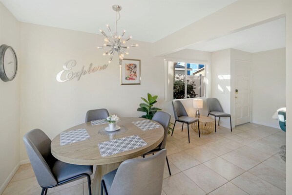 A dining area with a round table, grey chairs, a chandelier, and a wall-mounted clock.