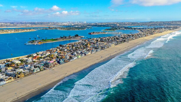 Luchtbeeld van een kustplaatsje met een lang strand, helderblauw water en een dichte verzameling gebouwen langs de kust.