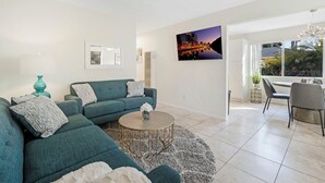 Een moderne woonkamer met een blauwgroene bank, een ronde salontafel en een televisiescherm met een stadsgezicht aan de muur.