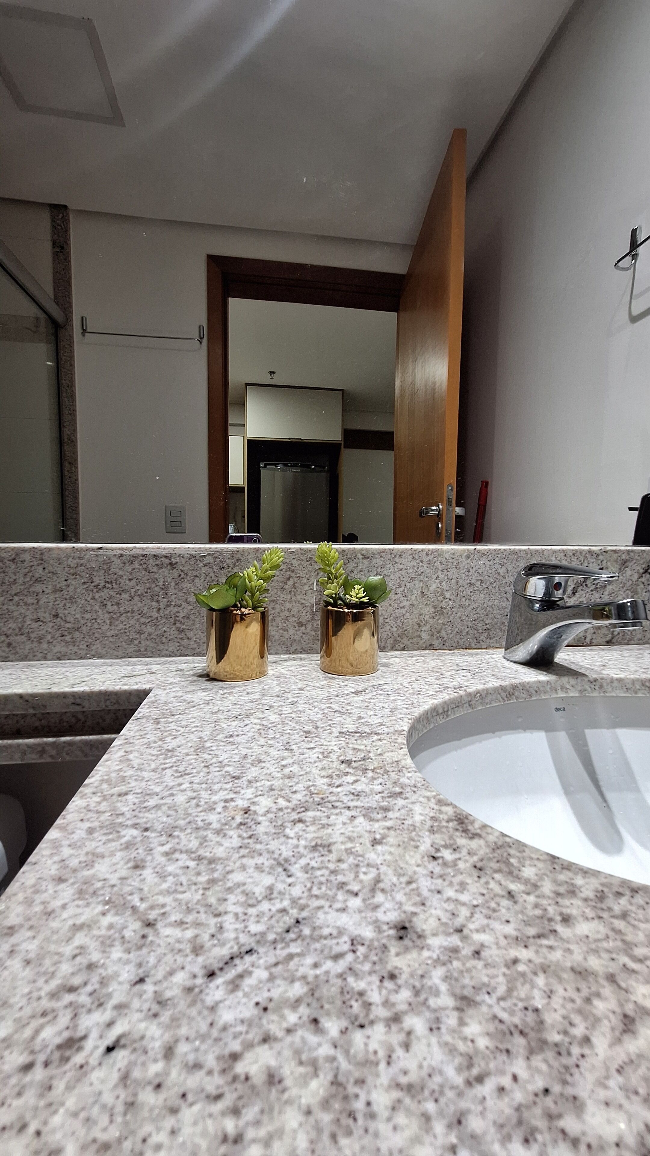 A bathroom with a granite countertop, two gold-colored plant pots, a sink, and a mirror reflecting a kitchen area.