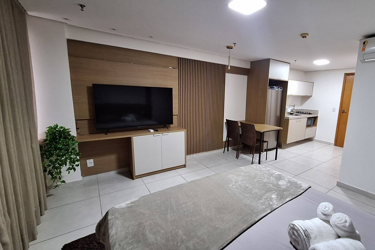 Una habitación de hotel moderna con televisión de pantalla plana, zona de comedor, kitchenette y cama con toallas.