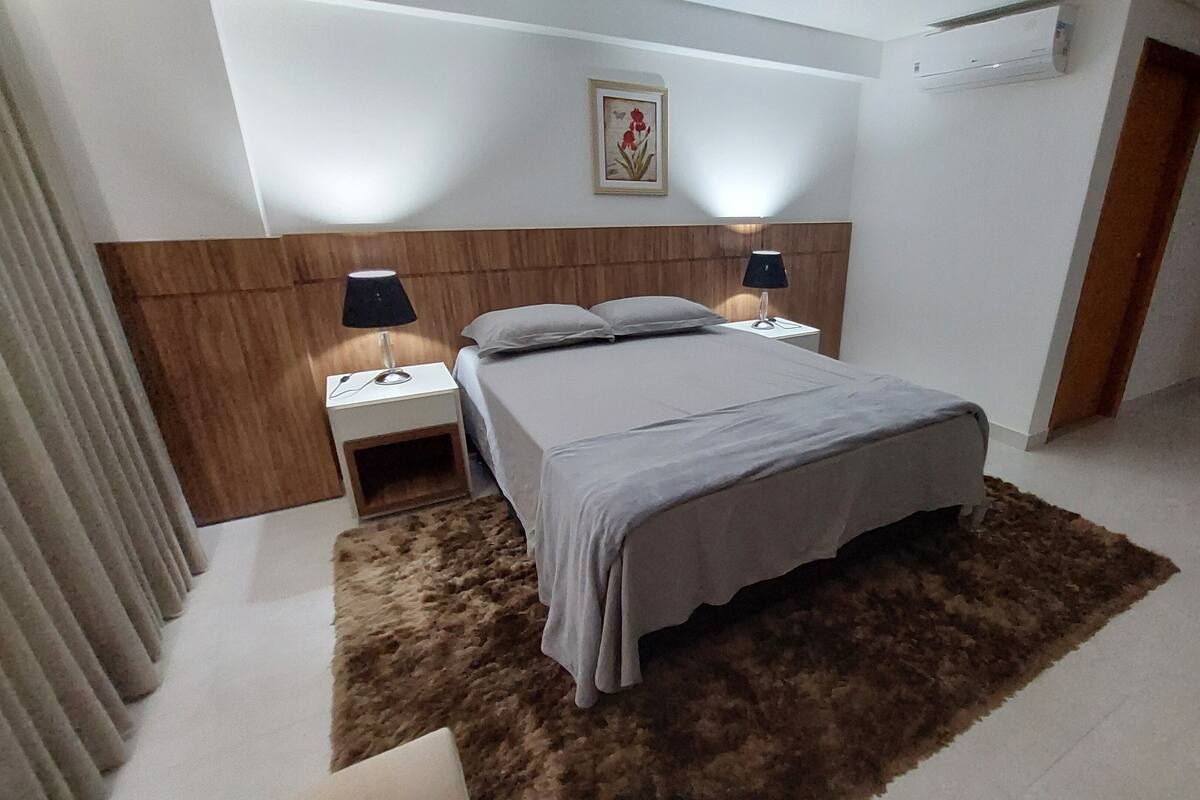Un dormitorio con cama, mesitas de noche, lámpara, un cuadro en la pared y un equipo de aire acondicionado.