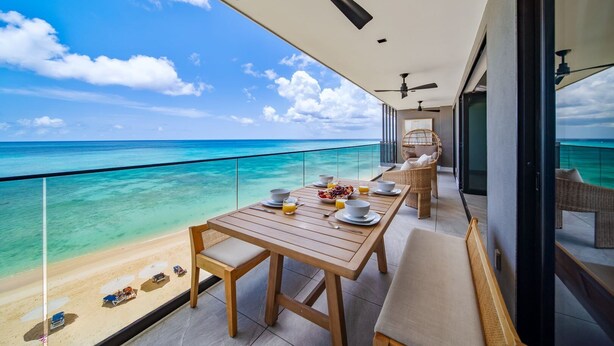 Un balcon avec une table en bois mise pour le petit-déjeuner, d’où l’on peut admirer une plage et une mer aux eaux turquoise.