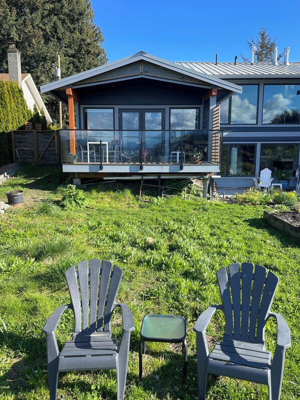 A modern house with large windows and a deck. There are two Adirondack chairs on the grass in front. - Endless views at Inlet Retreat! Relax in a centrally located beautiful studio! (Sechelt)