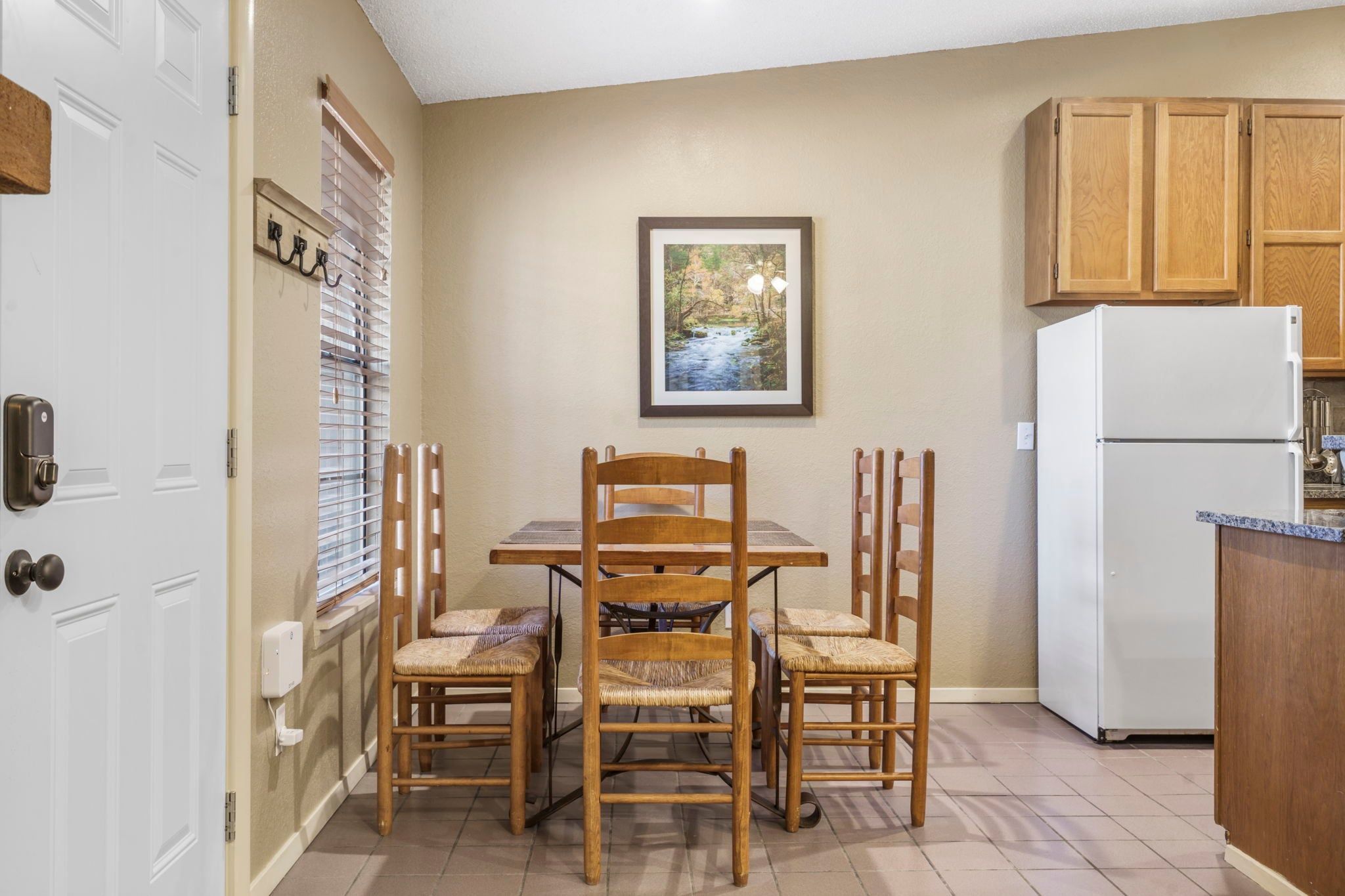 A kitchen with a dining table, chairs, a refrigerator, wooden cabinets, and a framed picture on the wall.