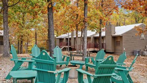 A campground with green Adirondack chairs arranged around a central fire pit, surrounded by trees with autumn foliage.