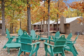 A campground with green Adirondack chairs arranged around a central fire pit, surrounded by trees with autumn foliage.