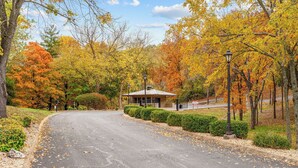 A paved road curving through a park with autumn foliage, a small building with a roof, and two street lamps.