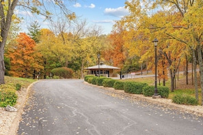 A paved road curving through a park with autumn foliage, a small building with a roof, and two street lamps.
