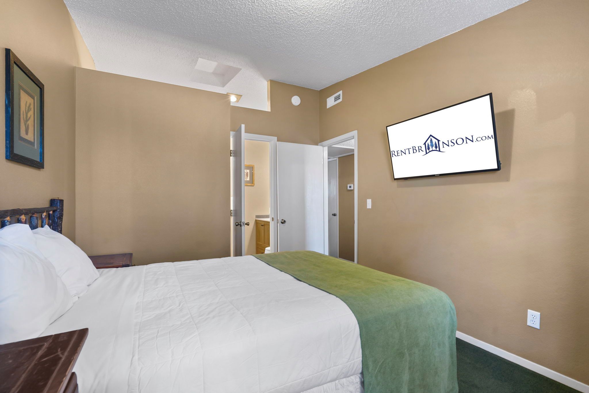 A hotel room with a bed, a green throw blanket, a flat-screen TV mounted on the wall, and a door leading to another room.