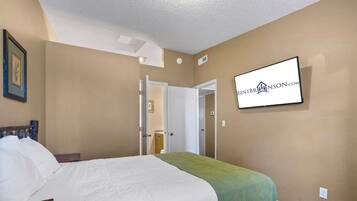 A hotel room with a bed, a green throw blanket, a flat-screen TV mounted on the wall, and a door leading to another room.